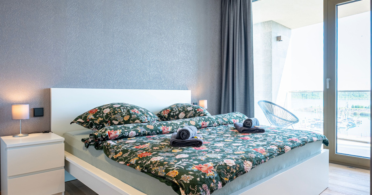 Blick in eines der Apartments von ALTUS.SLEEP: weißes Bett und Nachtschränke vor grauer Mustertapete, grüne Design-Bettwäsche, im Hintergrund bodentiefe Fenster mit Blick auf den See.