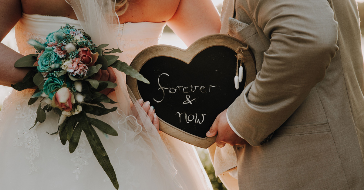 ein Brautpaar hält hinter sich den Brautstrauß sowie eine herzförmige Tafel mit der Aufschrift "forever & now" hoch.