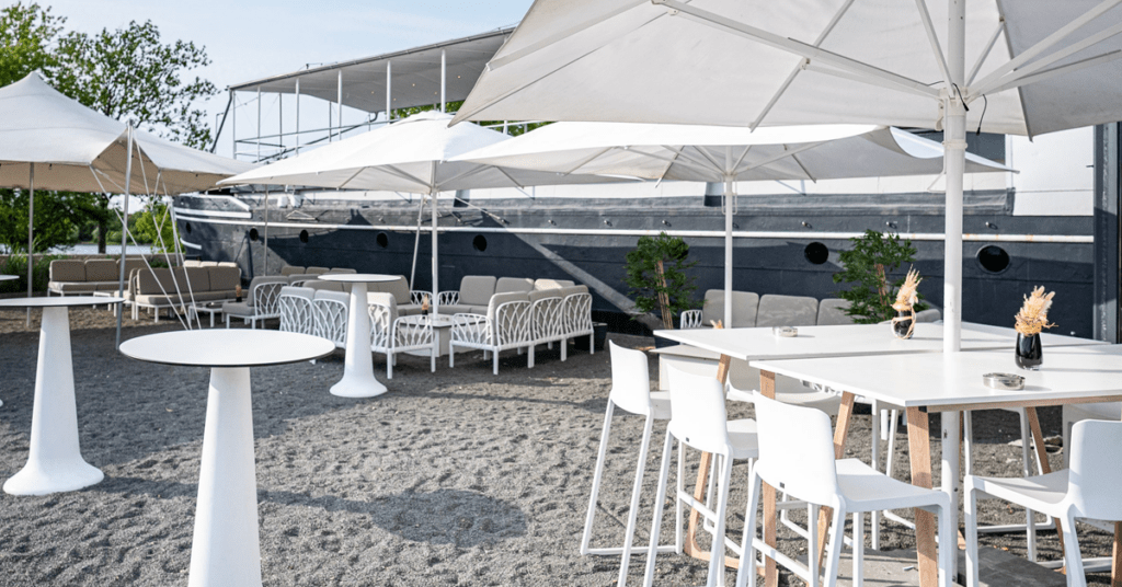 Das Bild zeigt den Außenbereich des amSee Leipzig mit weißen Möbeln und dem grauen Schiffsrumpf im Hintergrund. Für das Objekt wird ein Hausmeister gesucht.