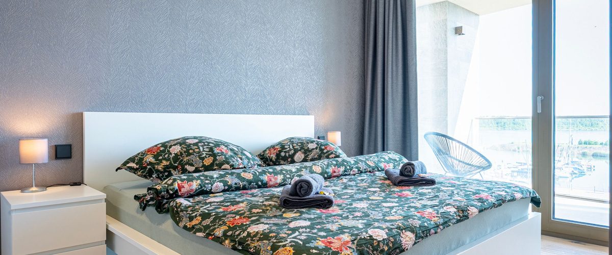 Blick in eines der Apartments von ALTUS.SLEEP: weißes Bett und Nachtschränke vor grauer Mustertapete, grüne Design-Bettwäsche, im Hintergrund bodentiefe Fenster mit Blick auf den See.