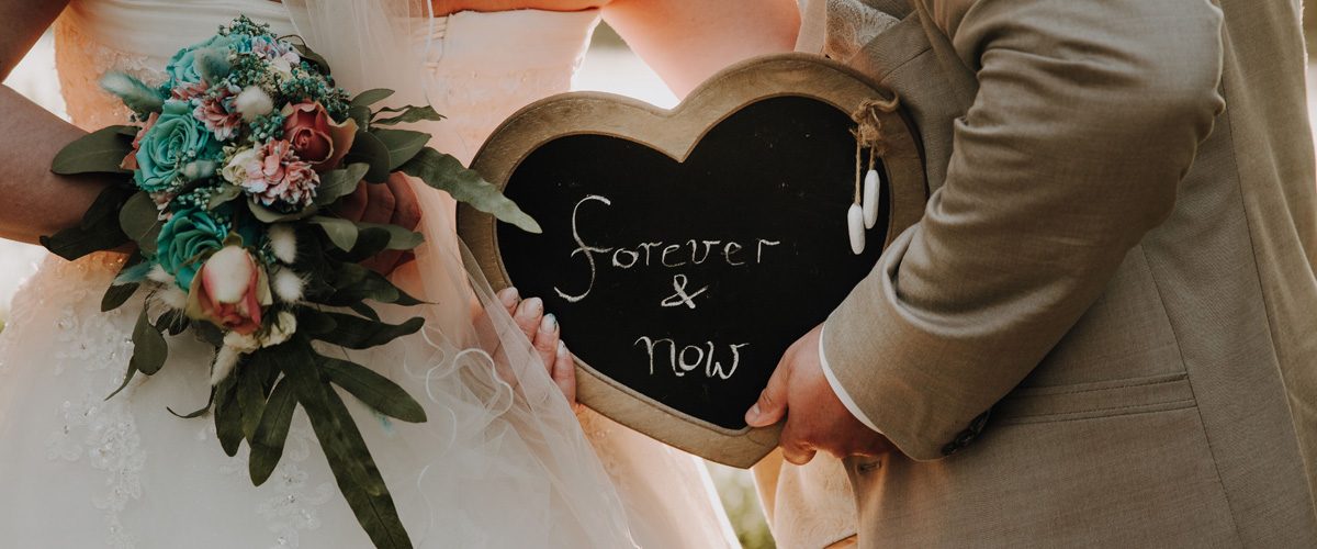 ein Brautpaar hält hinter sich den Brautstrauß sowie eine herzförmige Tafel mit der Aufschrift "forever & now" hoch.