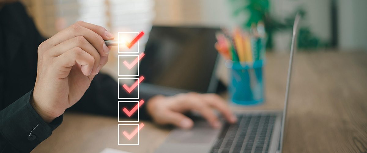 Stressfreie Eventplanung: Eine Person sitzt vor zwei Laptops an einem Tisch, im Vordergrund sind 5 Kästchen abgebildet (untereinander), die jeweils ein Häkchen enthalten. Das letzte Häkchen setzt die Person gerade mit einem stiftähnlichen Gegenstand in der Hand.